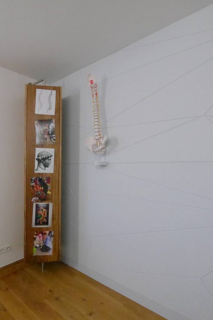 ein Eiche funriertes Drehrregal mit einer Türe auf der mit Magneten Bilder angebebracht sind. Daneben eine Wirbelsäule an der Wand aufgehöngt die mit Schallschluckendem Material verkleidet ist in IceBlue.