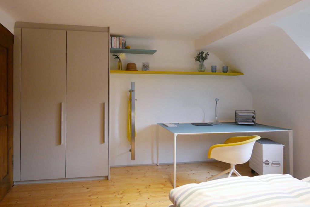 Raum in gelb, türis und hellgrau gehalten. Ein raumhoher Einbauschrank auf der linken Seite, farbige Regalbretter in die Schräge eingepasst und darunter ein Arper Schreibtisch in Kombination mit einer kleinen Bordbar als Bürocontainer.