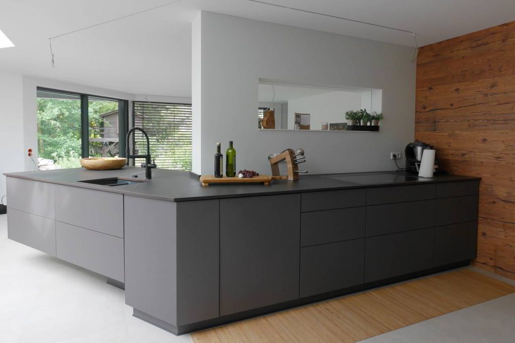 Antrazith farbene Küche, grifflos mit Keramik-Arbeitsplatte und flächenbündigem Kochfeld. Dahinter eine Wand mit einem Fenster in weiß gehalten.