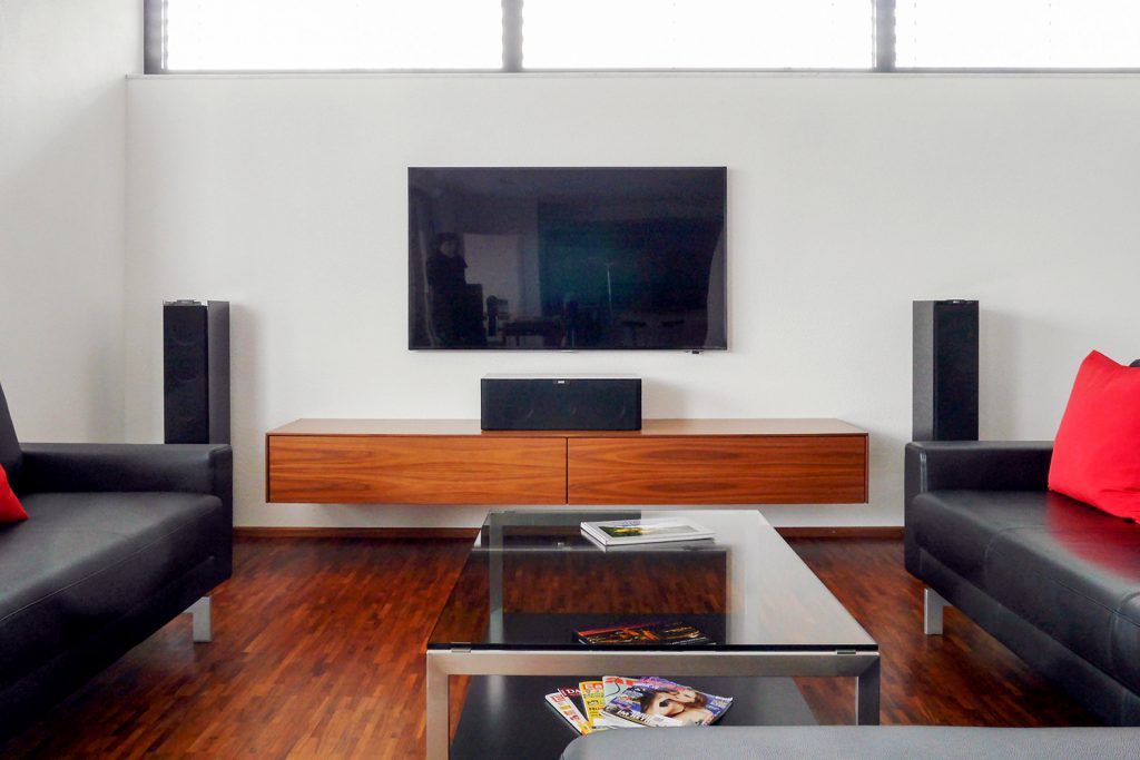 Wandhängendes TV-Sideboard mit einer Klappe und einer Schublade. Weiße Rückwand unter den Fenstern als schalldämmende Wand ausgebildet.