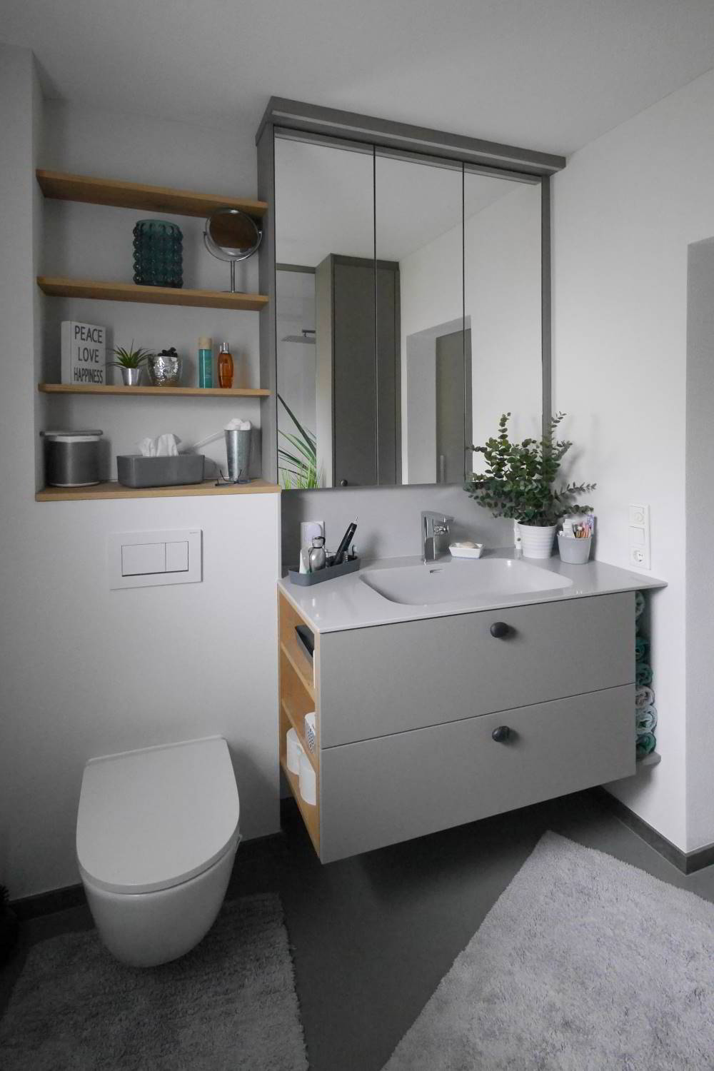 drei Regalböden aus Eiche furniert links über der Toilette angebracht. DAneben ein raumhoher Spiegelschrank mit direkter Beleuchtung. Darunter ein Waschtischunterschrank mit grauem Mineralgusswaschbecken und wandhängendem Waschtischunterschrank mit zwei Auszügen.