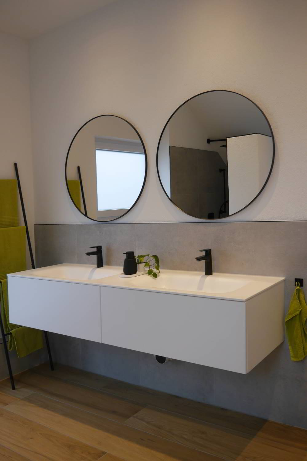 Badezimmer mit Waschtisch aus weiße Mineralguss. Darunter ein wandhängender weißer Waschtischunterschrank mit zwei Auszügen. Darüber sind zwei runde Spiegel angebracht.