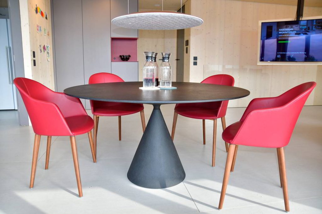 Hier steht ein schöner schwarzer Esstisch, rund in der Form mit einem Mittelfuß, der kegelförmig geformt ist. Oben drüber eine runde Lampe, das Nimbus Lighting Pad in hellgrau, diese wirkt fast schwebend, da sie nur mit drei Schnüren an der Decke hängt. Drum rum weinrote Leder bezogene Stühle.