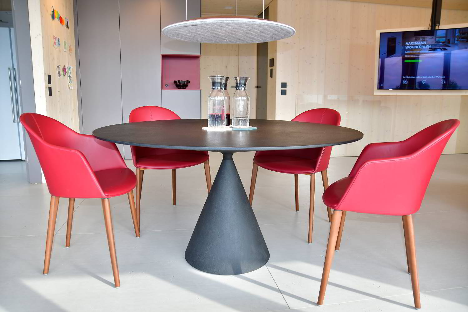 Hier steht ein schöner schwarzer Esstisch, rund in der Form mit einem Mittelfuß, der kegelförmig geformt ist. Oben drüber eine runde Lampe, das Nimbus Lighting Pad in hellgrau, diese wirkt fast schwebend, da sie nur mit drei Schnüren an der Decke hängt. Drum rum weinrote Leder bezogene Stühle.