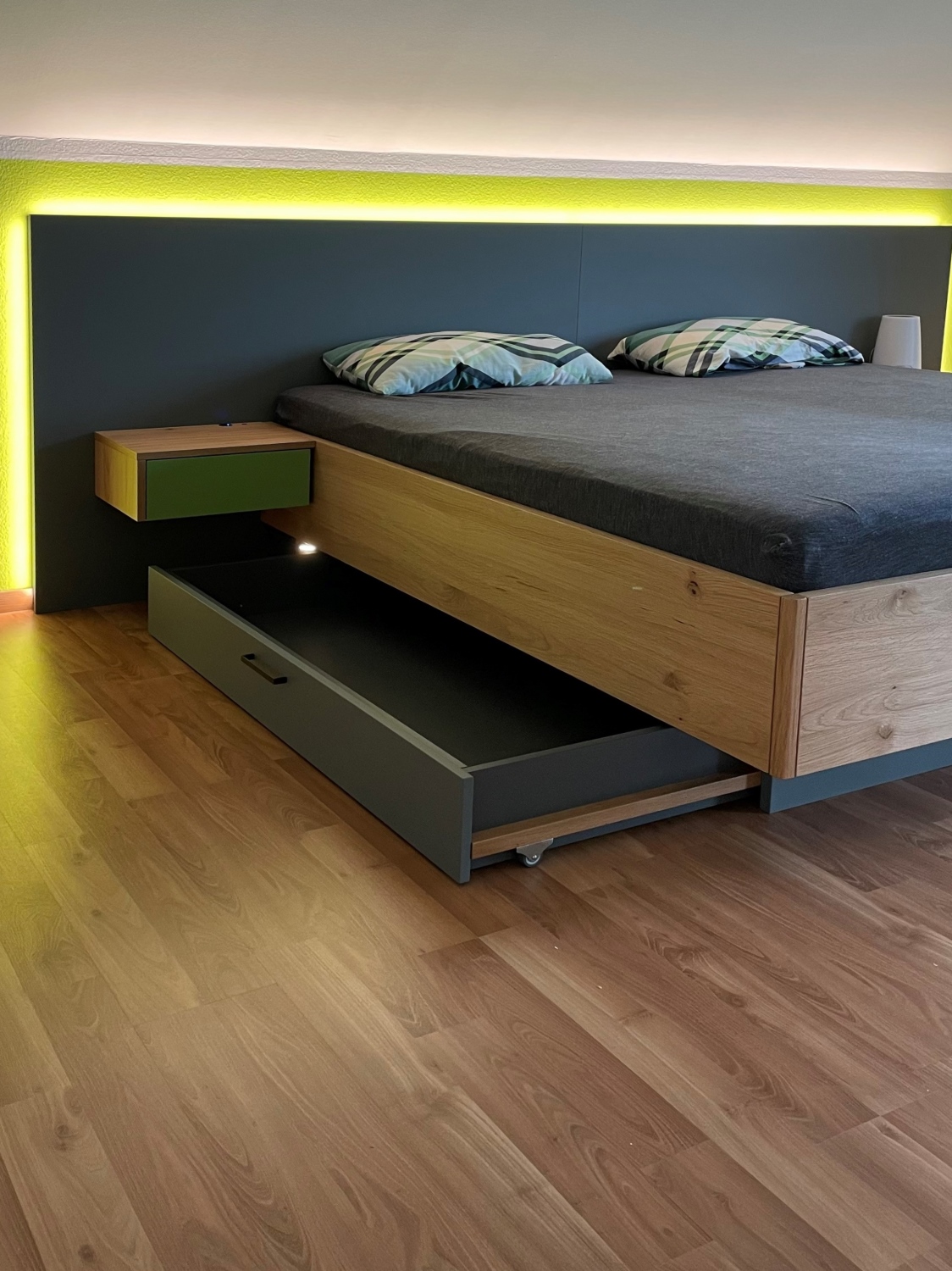 Ein Bett mit Holzumrandung, beleuchtetem schwarzen Kopfteil, einem hängendem Nachtisch mit einer grünen Front und einem ausgezogenen Schubkasten mit hoher Qualiät gefertigt von der Schreinerei Hartmann WOHNfühlen.