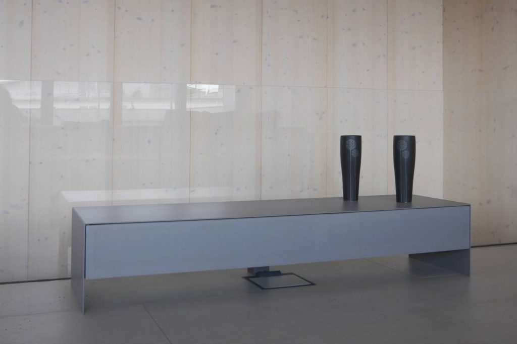 Sideboard in hellgrau mit dünner Abdeckplatte und Wangen aus Metall. Eine durchgängige lange Schublade vorne mit Lautsprecherstoff bezogen, farblich passend in hellgrau. Das Ganze steht in einem Raum vor einer Glaswand Richtung einer Treppe.