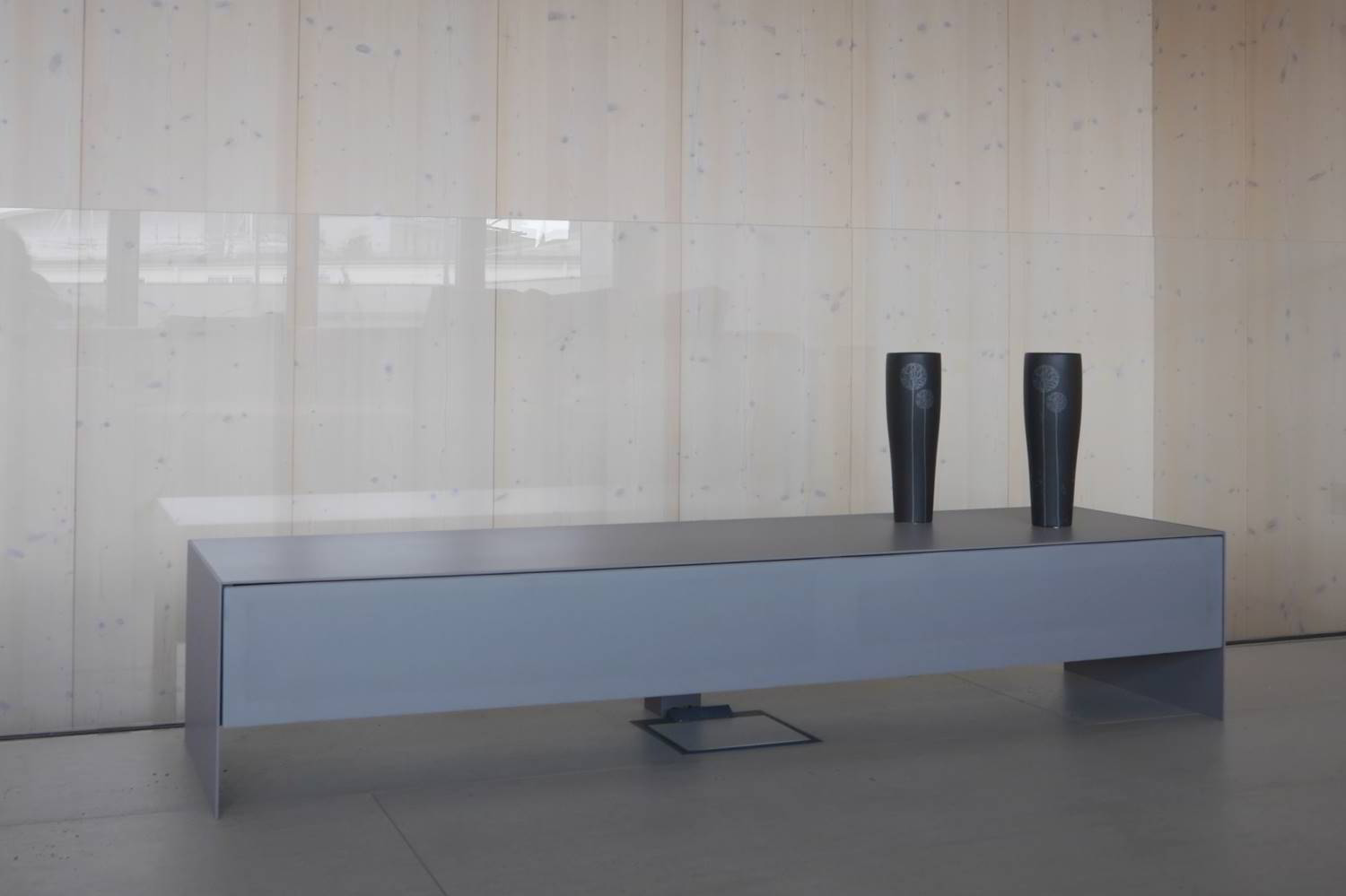 Sideboard in hellgrau mit dünner Abdeckplatte und Wangen aus Metall. Eine durchgängige lange Schublade vorne mit Lautsprecherstoff bezogen, farblich passend in hellgrau. Das Ganze steht in einem Raum vor einer Glaswand Richtung einer Treppe.