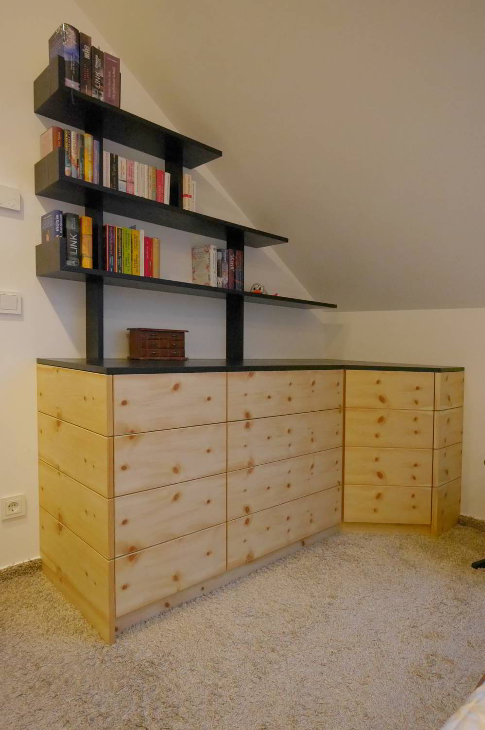 Sideboard über Eck laufend in Zirbe furniert, Abdeckplatte und aufgesetztes Regal in dunkel. Das ganze steht in einer Raumecke mit Dachschräge.