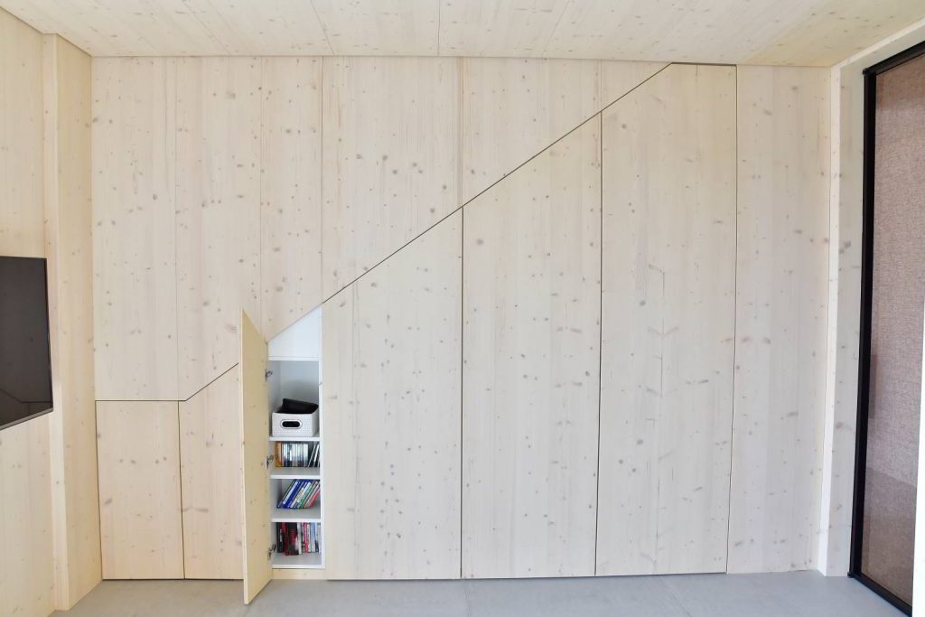 Wand in Fichte. Man sieht den Verlauf einer Treppe. Hier sind Türen integriert. Eine Türe steht leicht offen und weiße Fachböden vom Einbauschrank sind sichtbar.