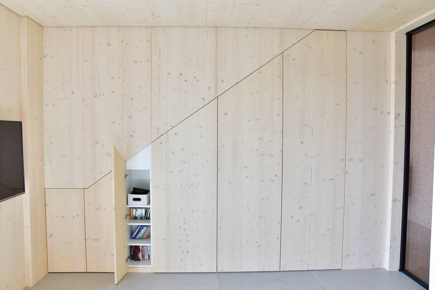 Wand in Fichte. Man sieht den Verlauf einer Treppe. Hier sind Türen integriert. Eine Türe steht leicht offen und weiße Fachböden vom Einbauschrank sind sichtbar.