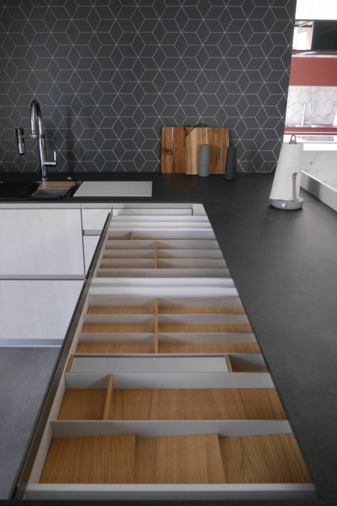 In einer weißen Küche sind mehrere Schubladen geöffnet, sie zeigen Besteckeinsätze mit Holzböden. Die schwarze Arbeitsplatte harmoniert mit der Rückwand, die ein elegantes weißes Muster trägt.
