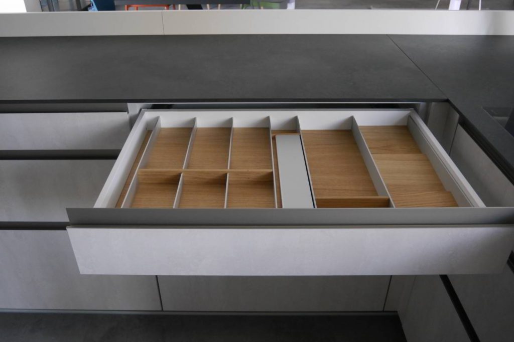 In einer hellen modernen Küche öffnet sich eine Schublade mit Griffmulde. Innen ein Besteckeinsatz und Holzboden. Küchenplanung bei Hartmann WOHNfühlen in Ellhofen.