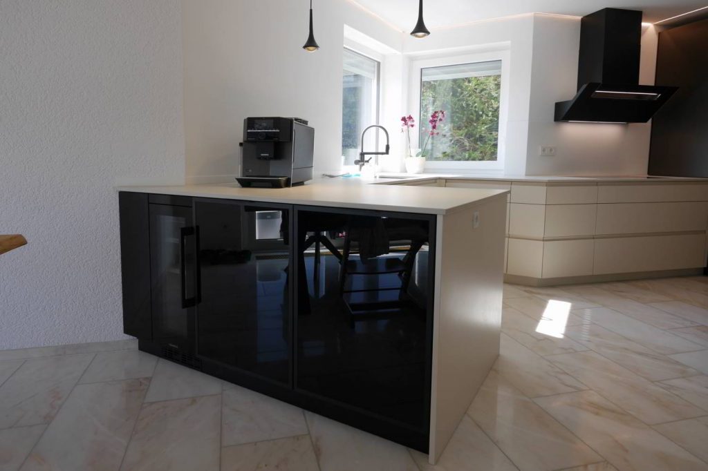 Küchentheke im Mittelpunkt ders Bildes, mit Fronten in Glas schwarz und links einem Weinkühlschrank. Helle Arbeitsfläche in Keramik und Boden.