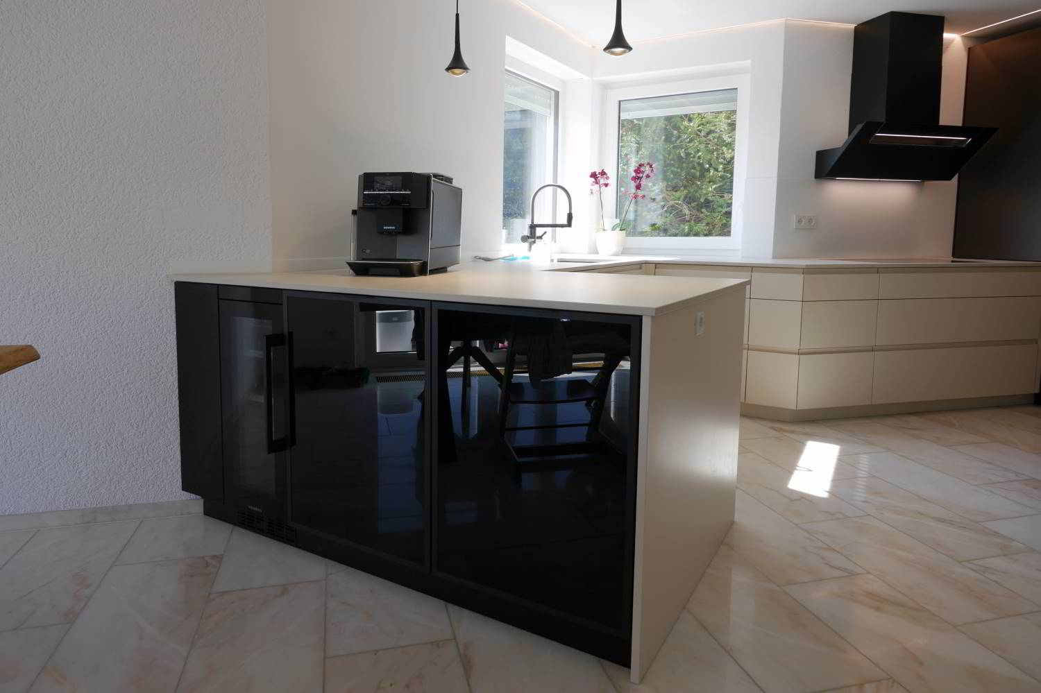 Küchentheke im Mittelpunkt ders Bildes, mit Fronten in Glas schwarz und links einem Weinkühlschrank. Helle Arbeitsfläche in Keramik und Boden.