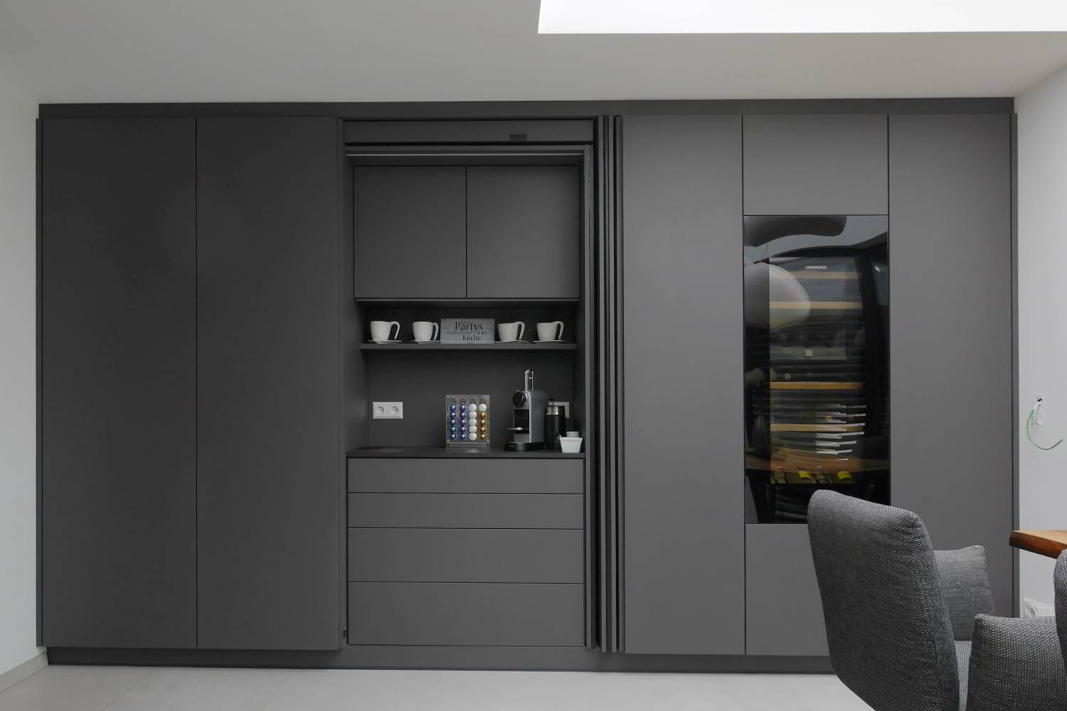 Hochschrankfront in Antrazit gehalten, mit Revego-Türe im linken Bereich integriert, die geöffnet ist. Hier ist die Kaffeebar zu sehen. Im Anschluss verschiedene Hochschränke und ein Weintemperierschrank. Alles bündig in die Wand eingebaut.