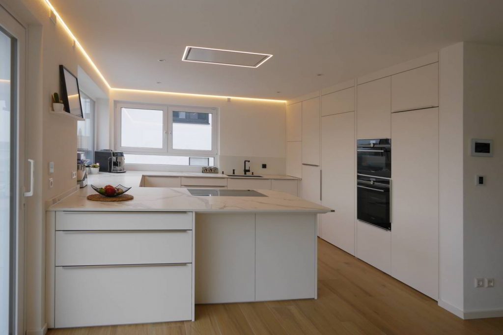 hell gehaltene Küche, mit flächenbündig in die Decke eingelassener Dunstabzugshaube von Novy. LED Beleuchtung in den Raumecken und Hochschränke bis an die Decke