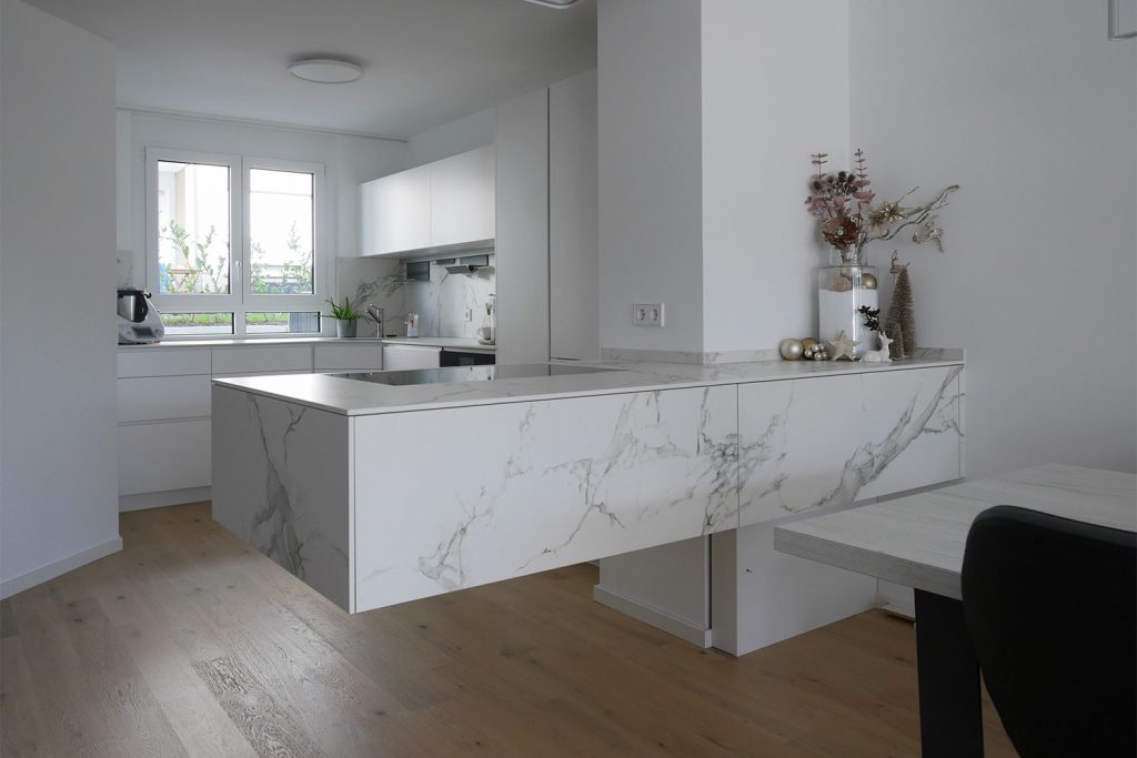 Offene, überwiegend in weiß gehaltene Einbauküche mit freihängendem Kochfeld, Keramikfronten und - platte. HartmannWOHNfühlen Ellhofen.