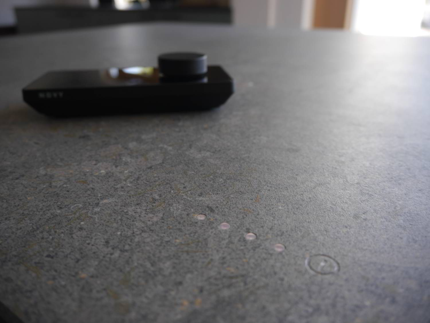 schwarzer Controller des Novy Undercover auf der dunkelgrau gefleckten Dekton Arbeitsplatte. Im Vordergrund der eingefräste An- und Ausschalter. DAneben die vier LED für die Anzeige der Kochzone.