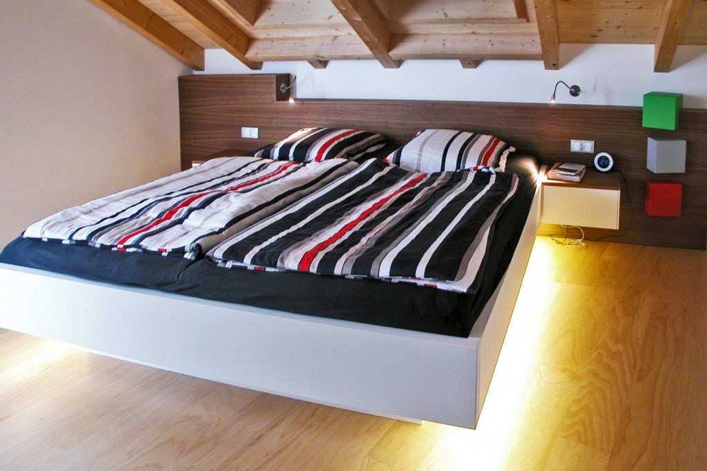 Freischwebende Bettanlage in Weißlack mit LED-Fußbodenbeleuchtung inklusive Bewegungsmelder. Kopfteil in Nussbaum Furnier mit Leseleuchten und drei kleinen, verdreht angeordneten Quadern. Hartmann WOHNfühlen, Ellhofen.