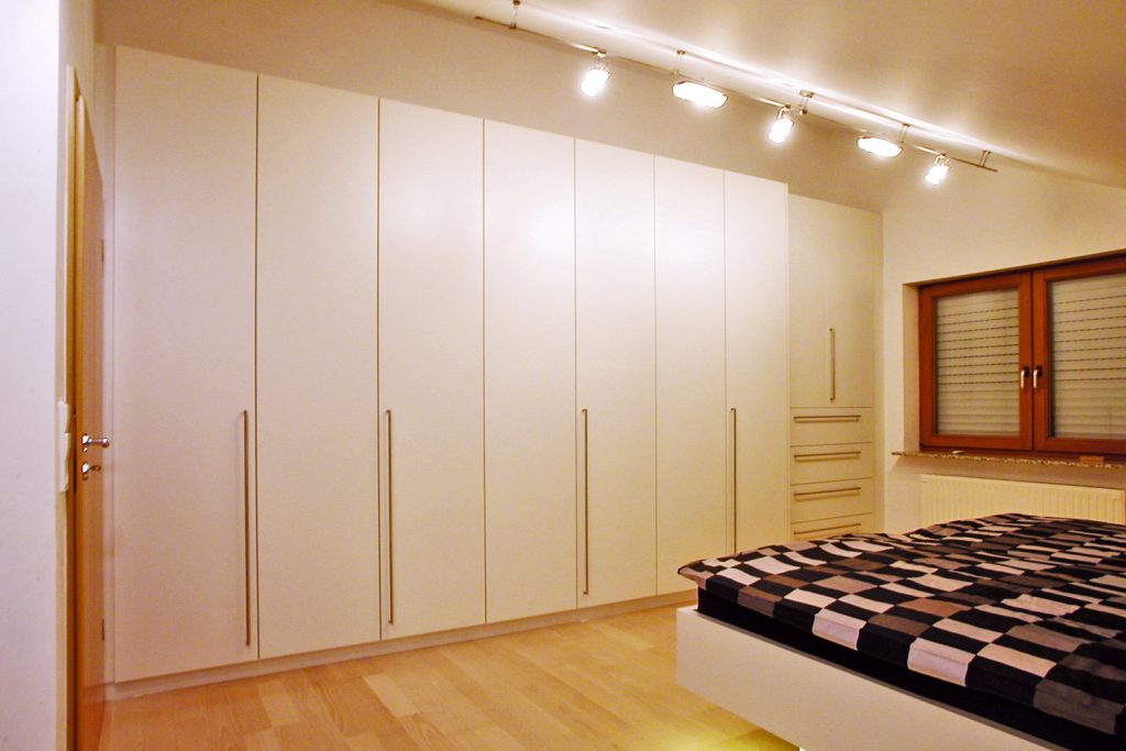 Großer in weiß lackierter Kleiderschrank mit zurückgesetztem Wäscheschrank. Griffleisten aus Metall. Schreinerei Hartmann, Ellhofen.