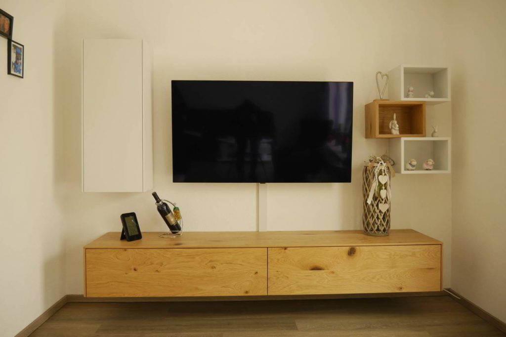 Vor eier weißen Wand hängendes Sideboard in Eiche furniert. Auf der linken Seite ein wandhängender Schrank mit einer Türe, rechts drei wandhängende Regale in Weiß und Eiche kombiniert. Mittige ein Fernsehr.