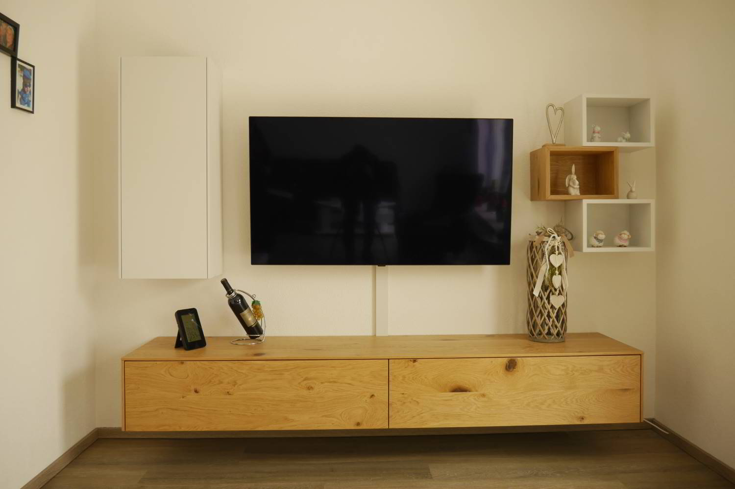 Vor eier weißen Wand hängendes Sideboard in Eiche furniert. Auf der linken Seite ein wandhängender Schrank mit einer Türe, rechts drei wandhängende Regale in Weiß und Eiche kombiniert. Mittige ein Fernsehr.