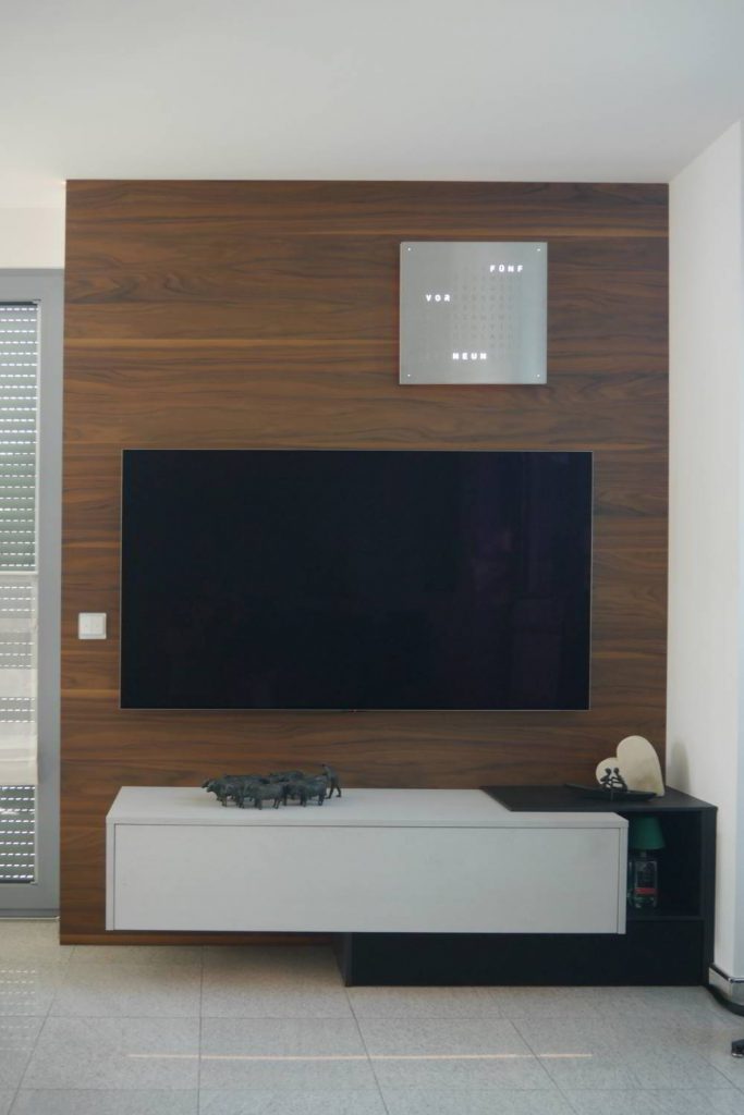 TV-Wand mit einer dunklen Rückwand in Nussbaumholz. Vorne ein wandhängendes Sideboard mit einer Klappe und ein weiterer Unterschrank mit Lautsprecherstoff ergänzt. Mittig ein Fernseher.