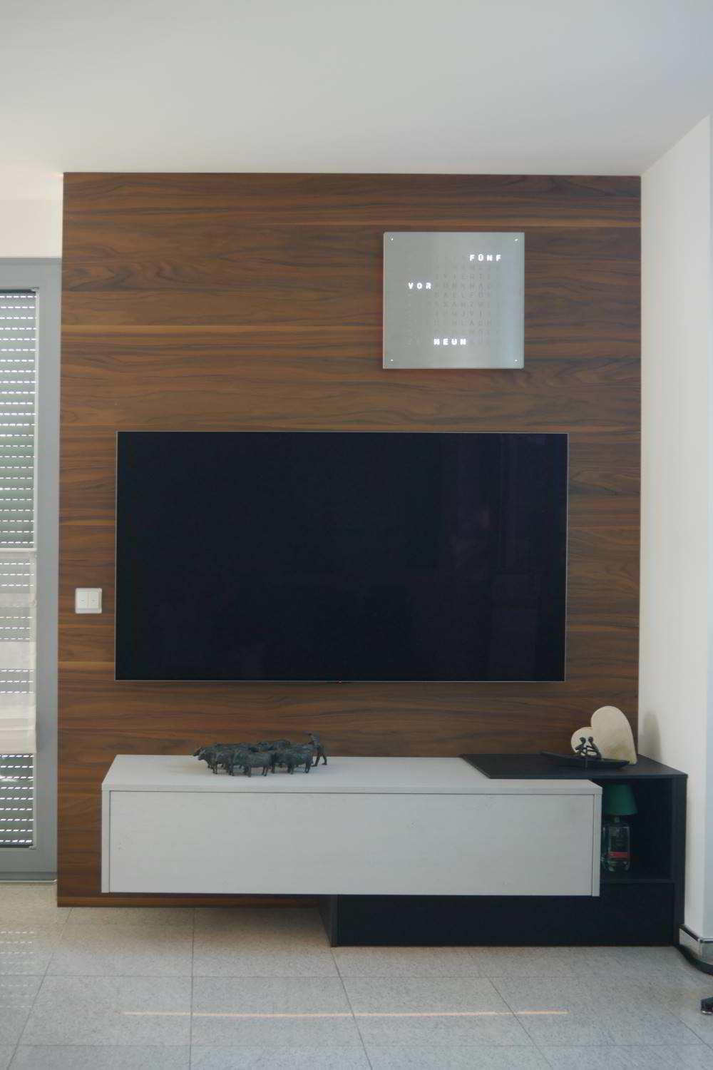 TV-Wand mit einer dunklen Rückwand in Nussbaumholz. Vorne ein wandhängendes Sideboard mit einer Klappe und ein weiterer Unterschrank mit Lautsprecherstoff ergänzt. Mittig ein Fernseher.