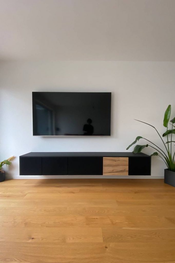weiße Wand mit Fernsehr und Pflanzen rechts und links. Fußboden aus Eiche. In der Mitte ein wandhängendes Sideboard mit 5 Klappen, davon 4 mit schwarzem Lautsprecherstoff bzw. Akustikstoff bezogen und eine in Eiche furniert. Abdeckplatten und Wangen in schwarz.