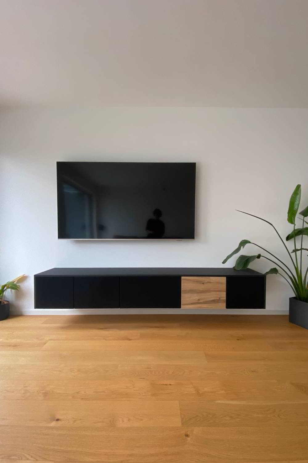 weiße Wand mit Fernsehr und Pflanzen rechts und links. Fußboden aus Eiche. In der Mitte ein wandhängendes Sideboard mit 5 Klappen, davon 4 mit schwarzem Lautsprecherstoff bzw. Akustikstoff bezogen und eine in Eiche furniert. Abdeckplatten und Wangen in schwarz.